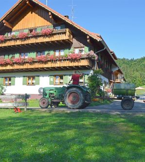 Ein landwirtschaftliches Gebäude mit blühenden Balkonen und einem grünen Traktor, der auf einem sonnigen Gelände steht. Im Hintergrund sind sanfte Hügel und Bäume zu sehen.