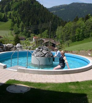 Ein runder Außenpool mit klarem Wasser, umgeben von Wiesen und Bergen. Im Hintergrund sind einige Bäume und Häuser sichtbar.