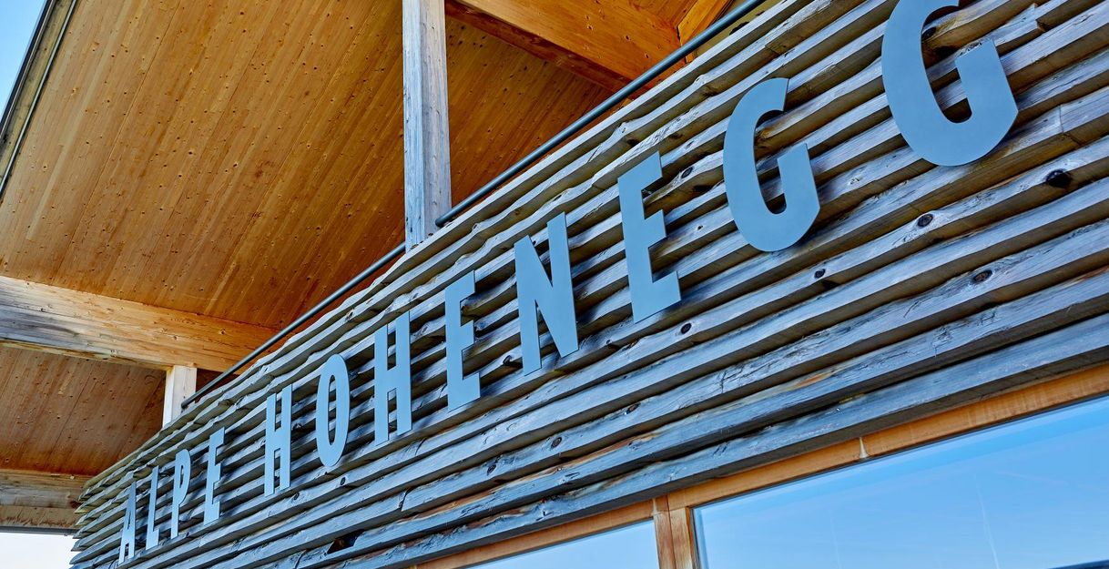 Ein modernes Gebäude mit einer Holzfassade und klaren Linien. Der Schriftzug „Hohenegg“ ist prominent an der Wand angebracht.