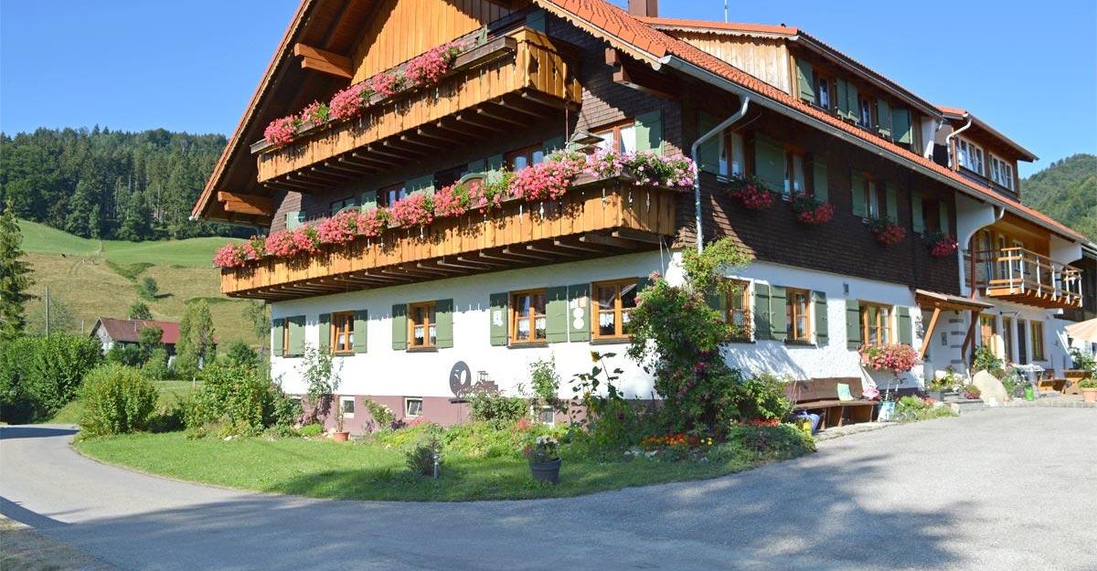 Ein traditionelles Haus mit schönem Holzbalkon und blühenden Pflanzen. Die Umgebung ist ländlich mit grünen Hügeln im Hintergrund.