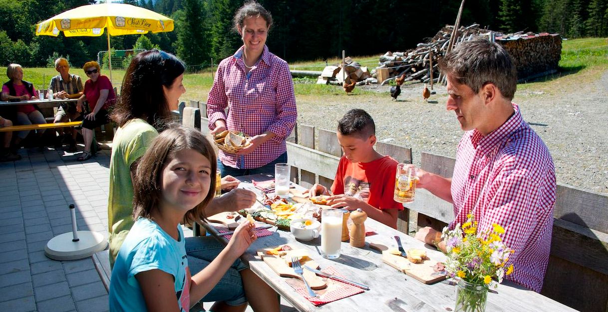 Familie isst auf der Lenzenalpe im Freien. Holztisch mit Brotzeit, Getränken und Blumen. Sonnenschirm spendet Schatten. Grüne Wiese und Bäume im Hintergrund.