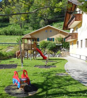 Ein Spielplatz mit einer Rutsche, Schaukeln und einer kleinen Kletteranlage in einem grünen Garten. Im Hintergrund steht ein Haus mit einem Balkon.