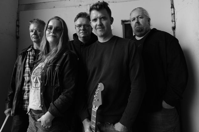 Schwarz-weiß-Bild der fünf Mitglieder mit Gitarre posierend vor einer Wand.