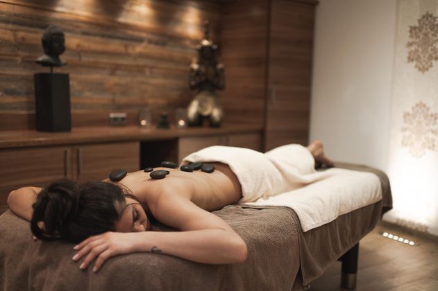 Eine entspannende Wellnessbehandlung mit einer Person, die auf einer Massagebank liegt. Steine sind auf ihrem Rücken platziert, und die Umgebung ist beruhigend gestaltet.