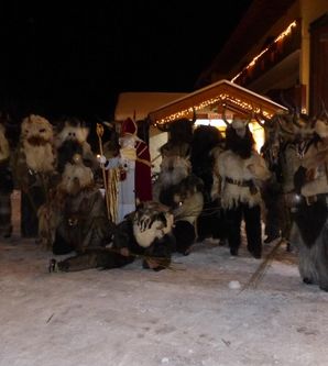 Eine Gruppe von verkleideten Personen in traditionellen Kostümen steht im Schnee. Im Hintergrund ist ein beleuchtetes Gebäude zu sehen.