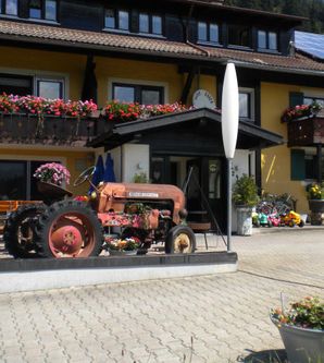 Ein roter Oldtimer-Traktor steht vor einem gelben Gebäude mit Blumenkästen. Das Haus hat Balkone und eine einladende Atmosphäre.