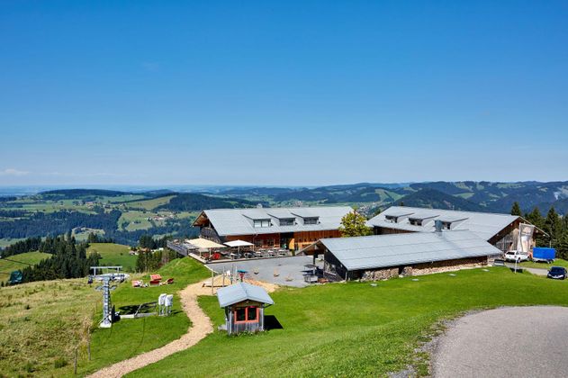 Eine schöne Berghütte mit einem weitläufigen Ausblick auf grüne Wiesen und Berge. Der Himmel ist klar und blau, ideal für einen Tag in der Natur.