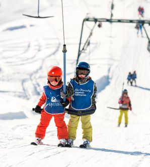 Zwei Kinder mit Skikleidung am Schlepplift, weitere Skifahrer im Hintergrund.