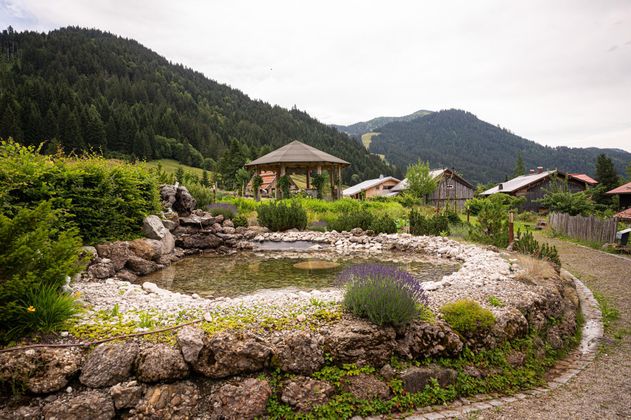 Runder Teich mit Steinufer und Lavendelbüschen, umgeben von üppigem Grün, Holzhäusern und bewaldeten Bergen unter bewölktem Himmel.