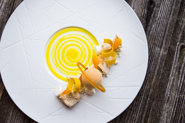 Ein kunstvoll angerichtetes Dessert auf einem weißen Teller, bestehend aus einer gelben Fruchtsauce und verschiedenen garnierenden Elementen. Dazu gehören eine Fruchteisportion, Orange- und Biskottestücke.