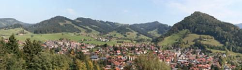 Eine malerische Stadt umgeben von sanften Hügeln und Bergen. Die grüne Landschaft und der klare Himmel schaffen eine idyllische Atmosphäre.