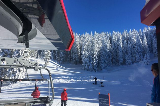 Blick von einer Talstation eines Sessellifts auf verschneite Piste mit Skifahrern, umgeben von tief verschneiten Bäumen