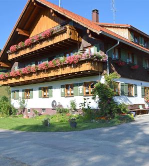 Ein traditionelles Haus mit schönem Holzbalkon und blühenden Pflanzen. Die Umgebung ist ländlich mit grünen Hügeln im Hintergrund.