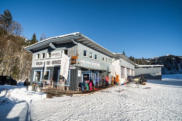 Gebäude von Planet B. Skischule und Verleih in Balderschwang. Graue Holzfassade, weiße Fensterrahmen. Schnee liegt. Blauer Himmel.