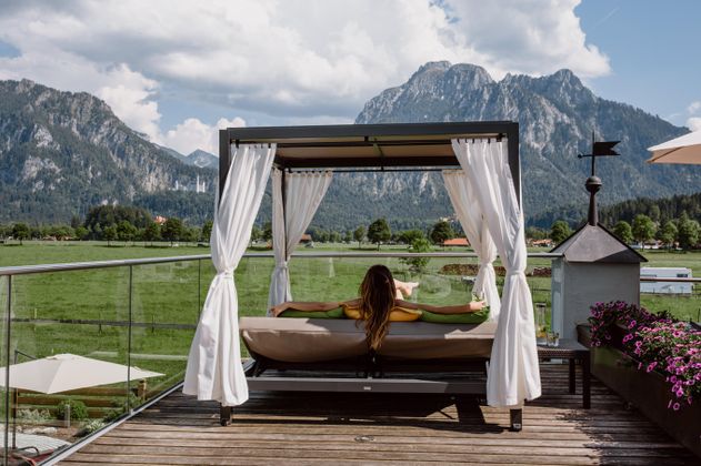 Eine entspannte Person liegt auf einer Liege mit Blick auf die Berge. Die Umgebung ist grün und einladend, mit weißen Vorhängen und blühenden Pflanzen.
