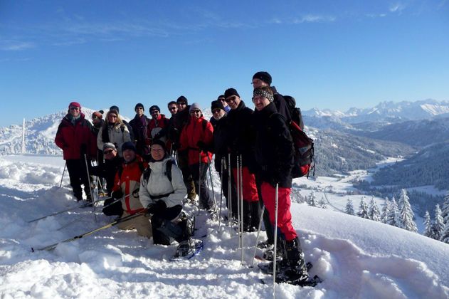 Gruppe von etwa 15 Personen mit Schneeschuhen und Skistöcken im Schnee. Bunte Winterkleidung. Verschneite Landschaft und Berge im Hintergrund. Blauer Himmel.