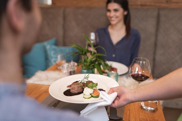 Ein schön angerichtetes Gericht wird einem Gast in einem gemütlichen Restaurant serviert. Im Hintergrund sitzt eine lächelnde Frau mit einem Glas Wein.