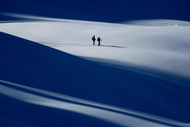 Zwei dunkle Silhouetten von Personen beim Skilanglauf in einer weiten, verschneiten Landschaft. Lange Schatten auf dem Schnee. Blauer Himmel.