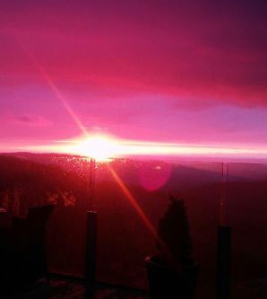 Ein atemberaubender Sonnenuntergang mit leuchtenden rosa und lila Farben über der Berglandschaft. Die Aussicht ist von einem Balkon mit einer Kante aus Glas umgeben.
