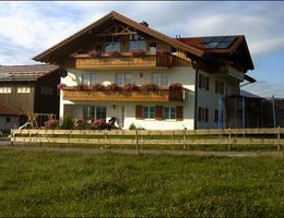 A traditional Bavarian house with several balconies and flowers. The building is situated in a rural setting with meadows and a hangar in the background.