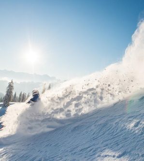 Ein Skifahrer in blauem Anzug fährt steilen Tiefschnee Hang hinab und wirbelt Schnee auf.