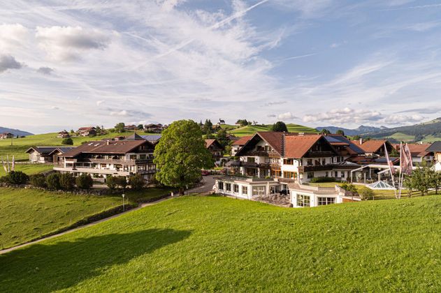 Eine malerische Landschaft mit gepflegten Wiesen und traditionellen Gebäuden. Im Hintergrund erstrecken sich Berge und ein klarer Himmel.