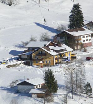 Eine ruhige Winterlandschaft mit verschneiten Hügeln und mehreren Häusern. Die Gebäude sind umgeben von schneebedeckten Bäumen und einem klaren Blau am Himmel.