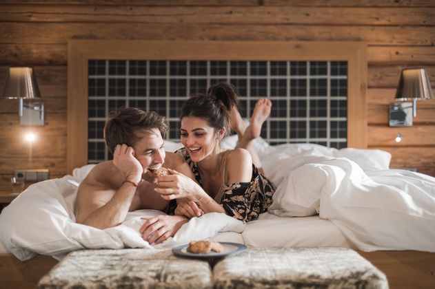 Ein glückliches Paar liegt lächelnd im Bett und genießt einen tollen Moment. Um sie herum sind weiche Kissen und eine gemütliche Holzverkleidung.