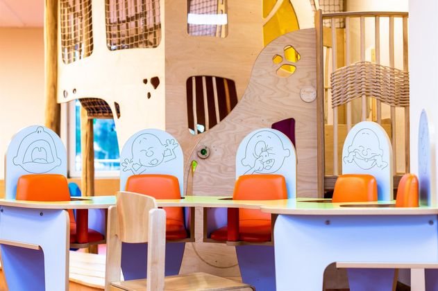 Ein farbenfroher Spielbereich mit einem Spieltrum im Hintergrund und einem Tisch mit bunten Stühlen. Die Stühle sind mit lustigen Gesichtern verziert, die Kinder ansprechen.